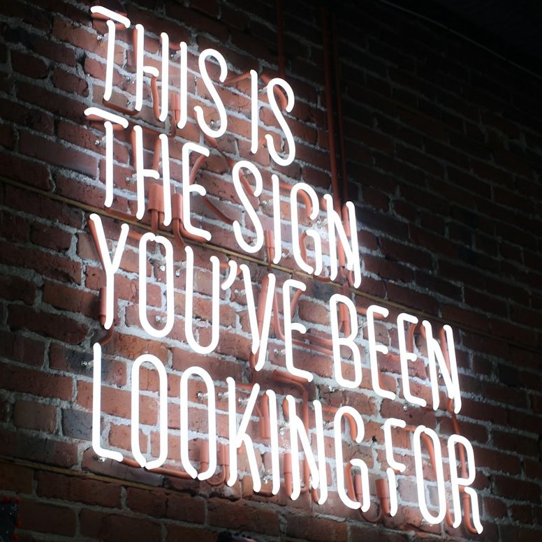 Texte en néon sur mur en brique : "Ceci est le signe que vous cherchiez."